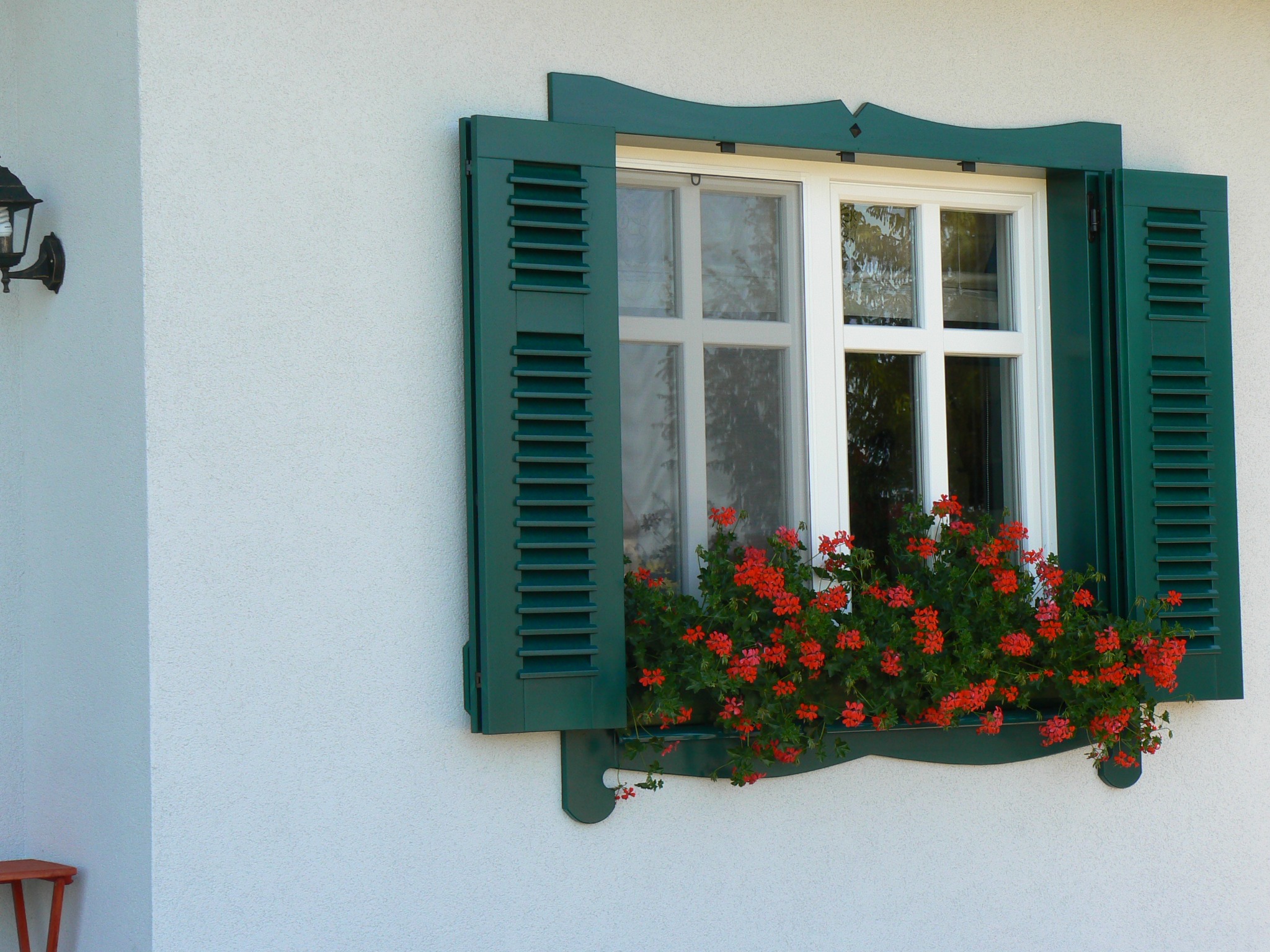 Białe okno z zielonymi okiennicami i skrzynką z czerwonymi pelargoniami na tle białej ściany.
