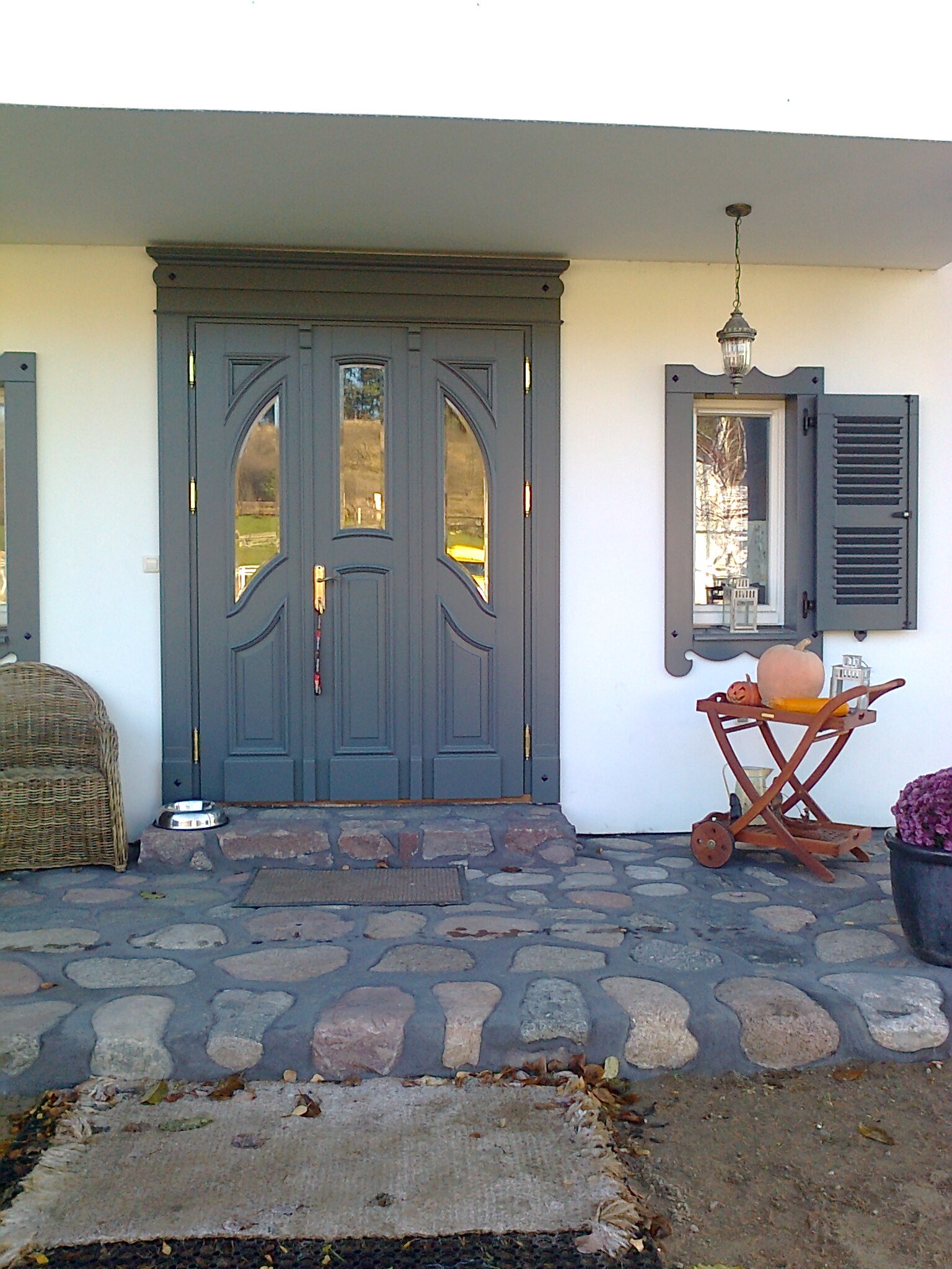 Szare drzwi wejściowe z szybami i dekoracyjnym obramowaniem, uzupełnione o okno z okiennicami, lampę wiszącą i wózek z dyniami na kamiennym chodniku.