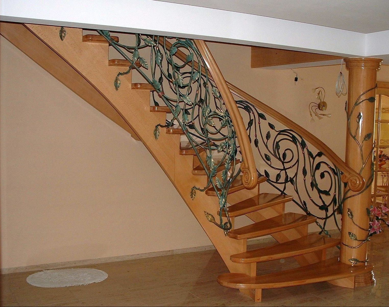 Drewniane schody z ozdobną, metalową balustradą z motywem roślinnym, zakończoną spiralnym elementem, wsparte na kolumnie z podobnym dekorem, w jasnym wnętrzu.