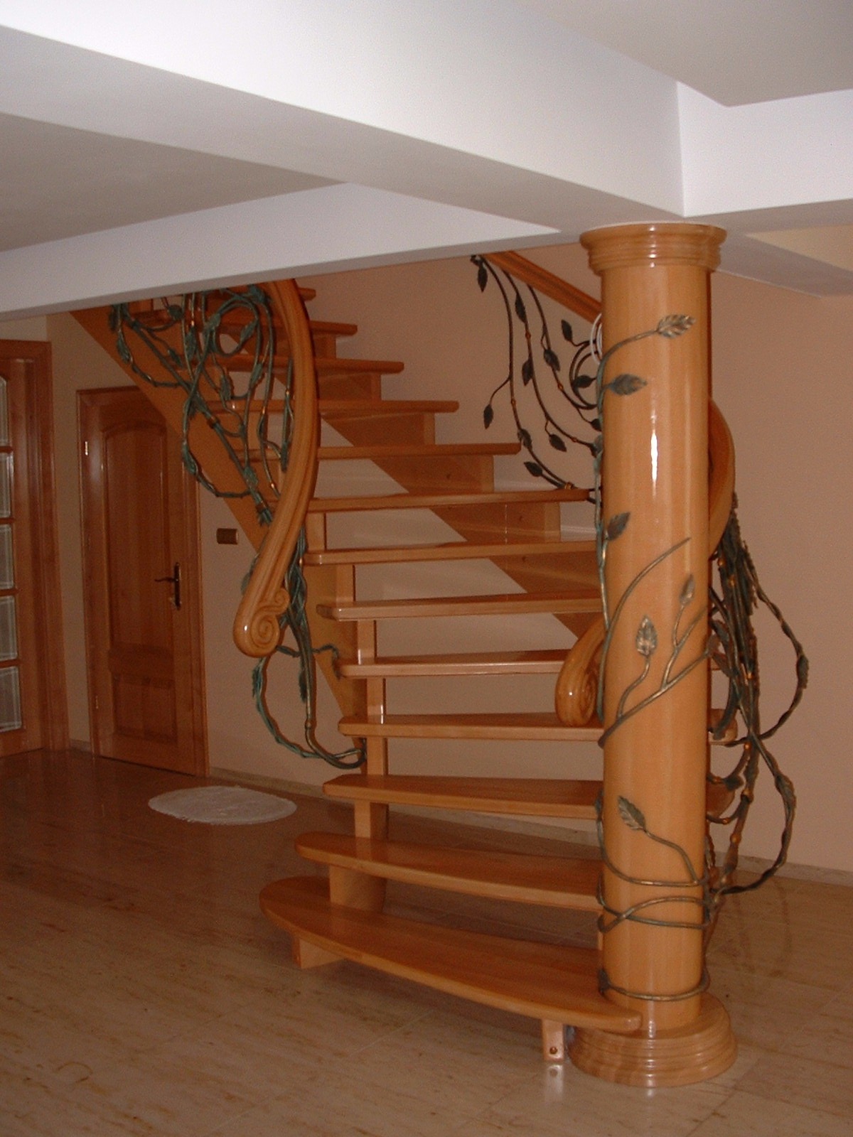 Drewniane spiralne schody z dekoracyjną metalową balustradą w kształcie pnączy z liśćmi, wsparte na centralnym słupie również oplecionym metalowymi elementami, widoczne drzwi w tle.