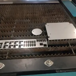 Metalowe elementy o różnych kształtach (okrąg, prostokąt z wycięciami, kwadrat z otworami) leżą na stole roboczym maszyny do obróbki metali.