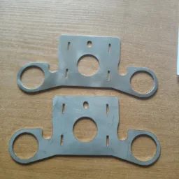 Dwie identyczne, płaskie, metalowe płytki z wyciętymi otworami (okrągły centralny otwór i dwa okrągłe otwory na końcach ramion) oraz podłużnymi nacięciami, leżące na drewnianym blacie.