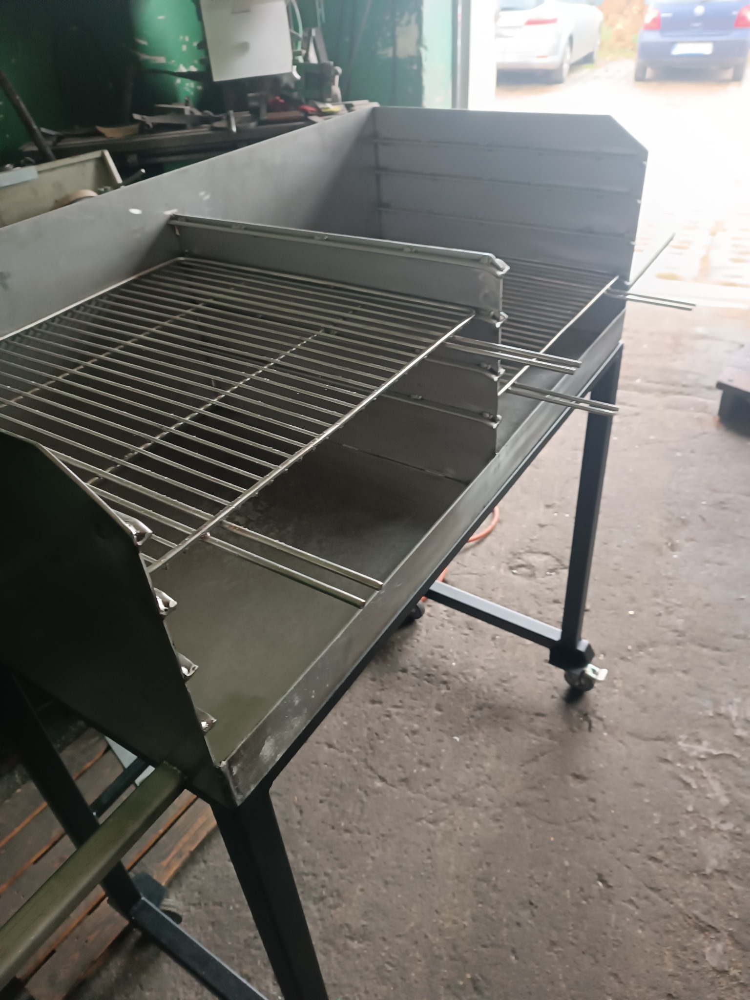 Metalowy grill na kółkach z rusztem, wykonany ze stali, w warsztacie z widocznymi śladami użytkowania na podłodze.