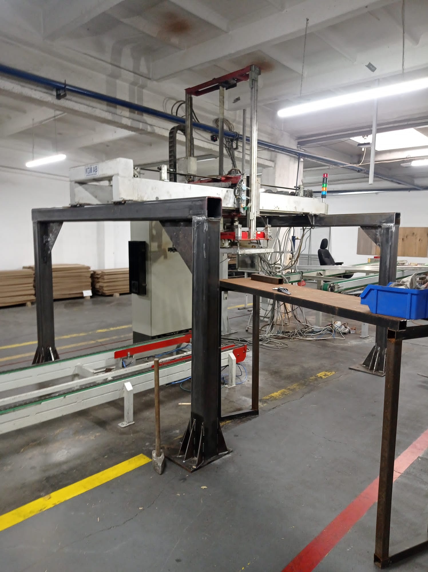 Wnętrze hali produkcyjnej z maszyną CNC, metalową konstrukcją ramową, stołem roboczym z niebieskim pojemnikiem i narzędziami, oraz paletami materiałów w tle.