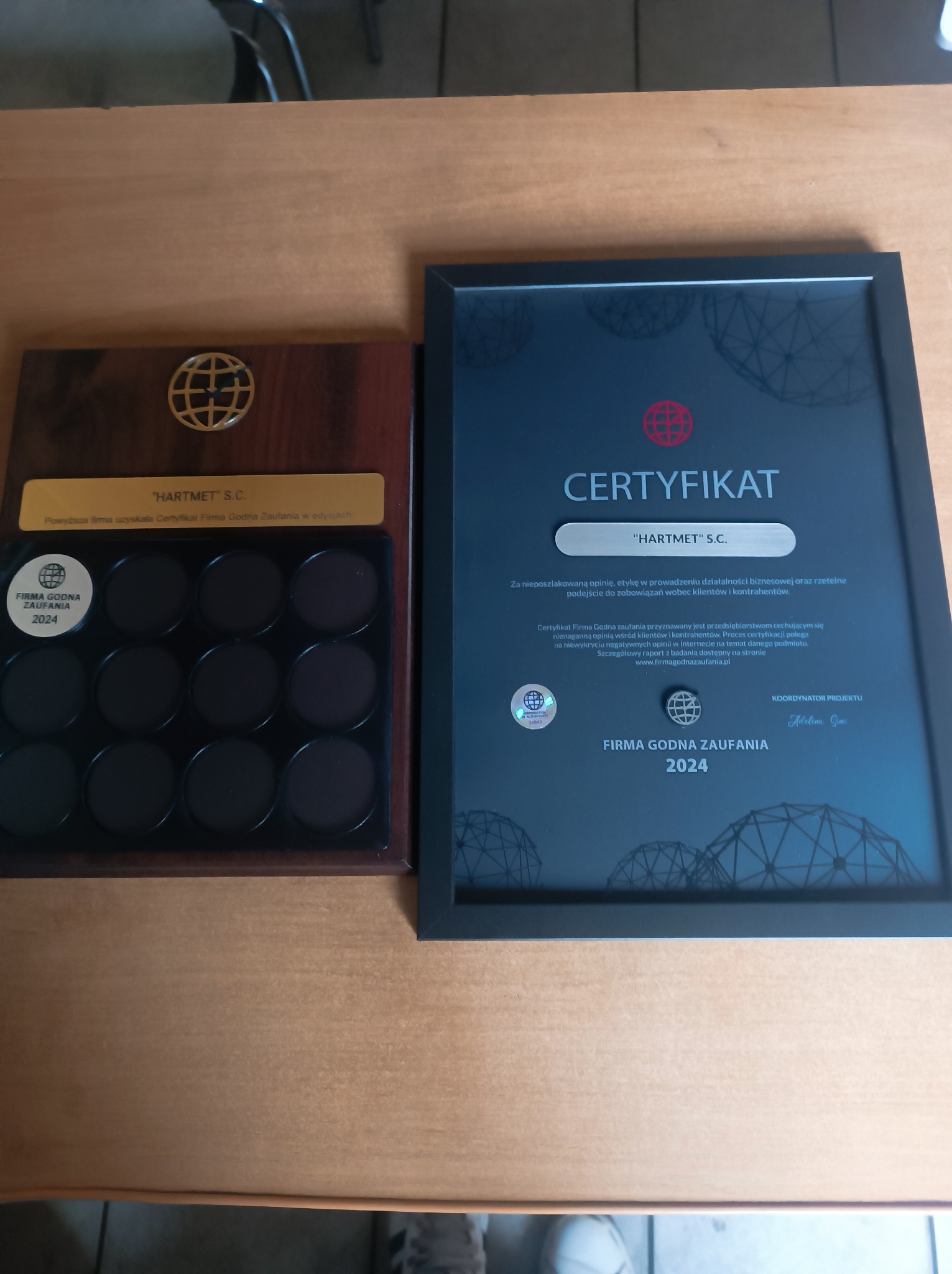 Certyfikat 'Firma Godna Zaufania 2024' dla 'HARTMET S.C.' w czarnej ramce, obok drewniana tabliczka z logo i okrągłymi polami na próbki lub odlewy.