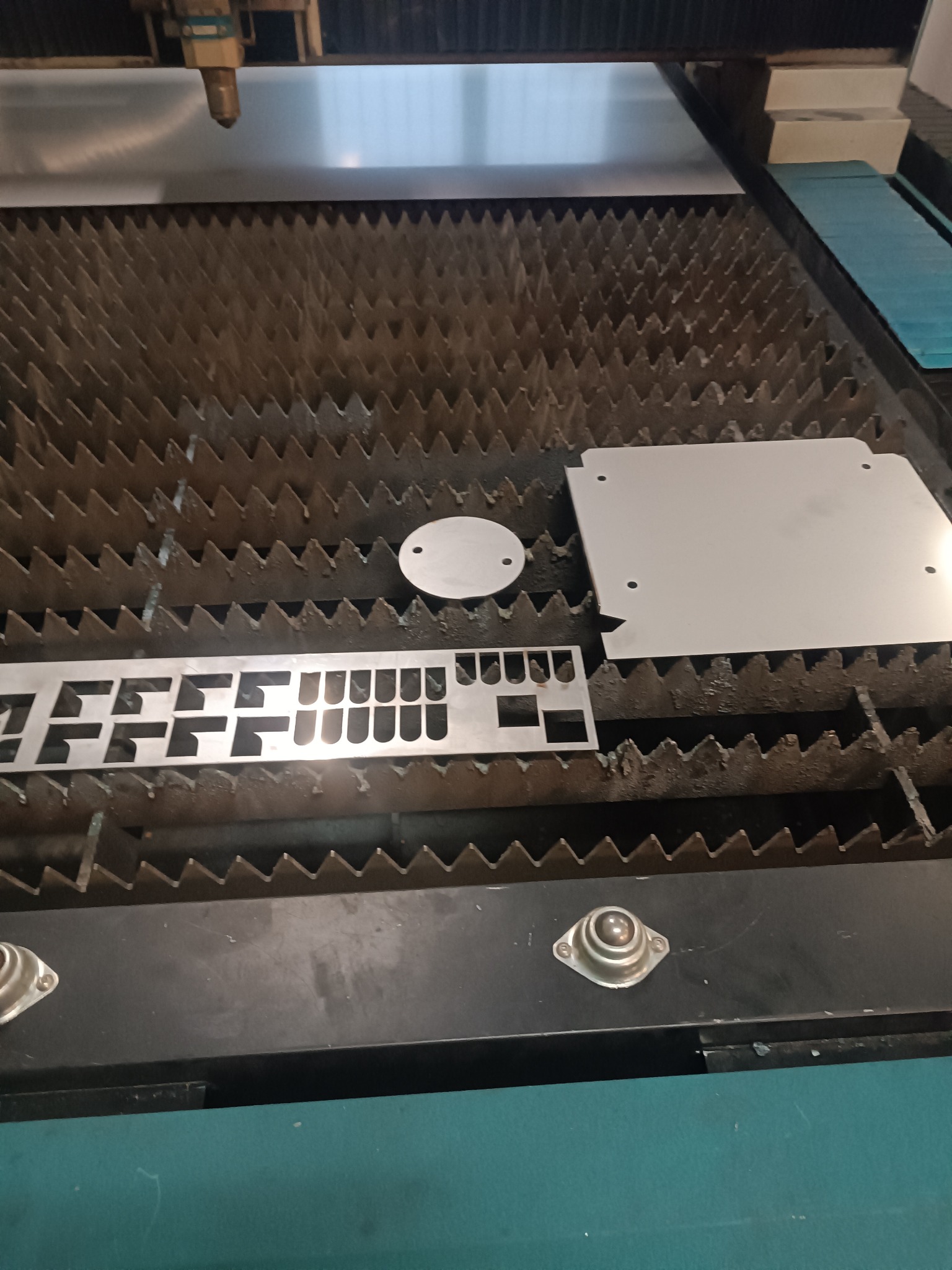 Metalowe elementy o różnych kształtach (okrąg, prostokąt z wycięciami, kwadrat z otworami) leżą na stole roboczym maszyny do obróbki metali.