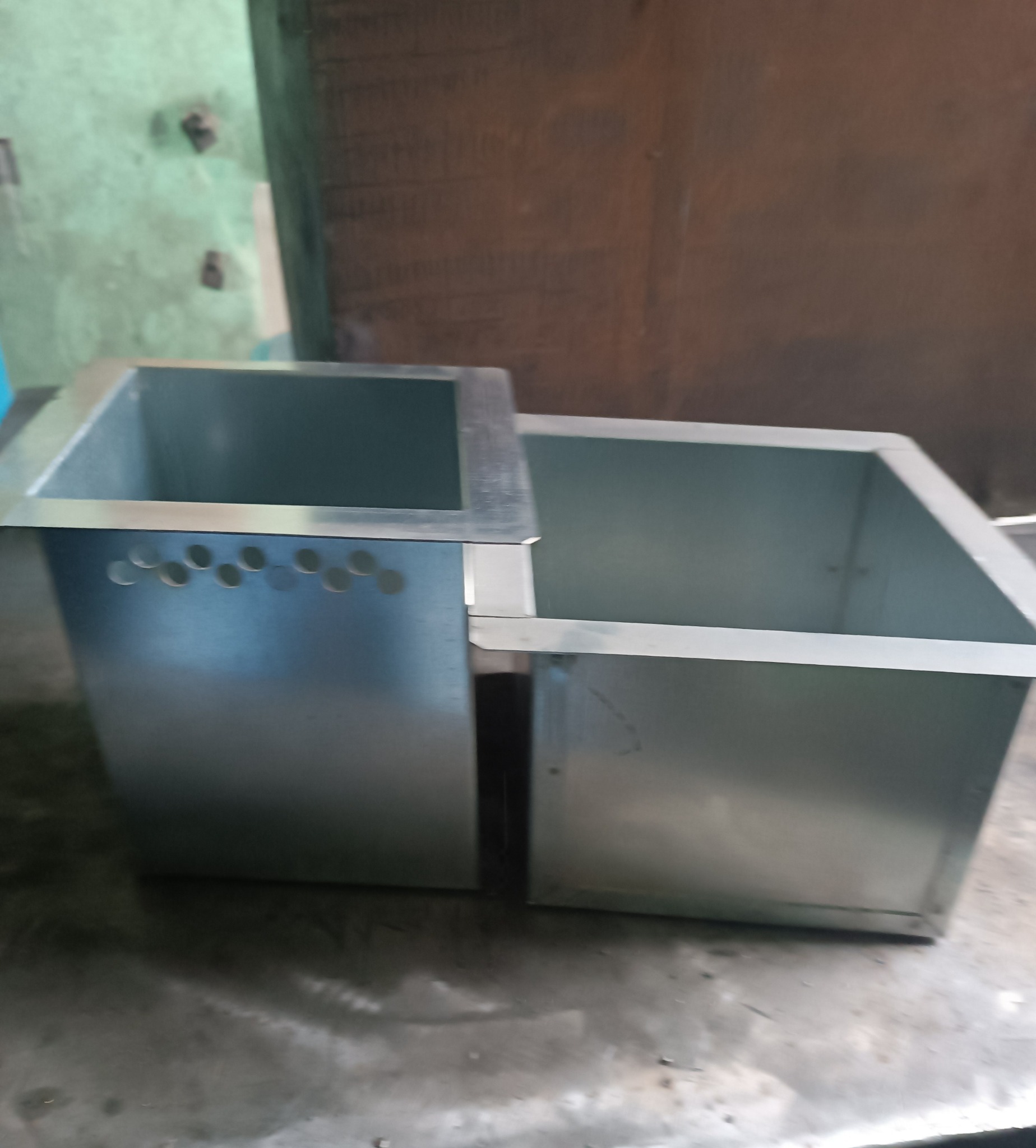 Dwa połączone, kwadratowe, metalowe pojemniki, jeden z rzędem okrągłych otworów wentylacyjnych w dolnej części ściany bocznej, na szarej, metalowej powierzchni.