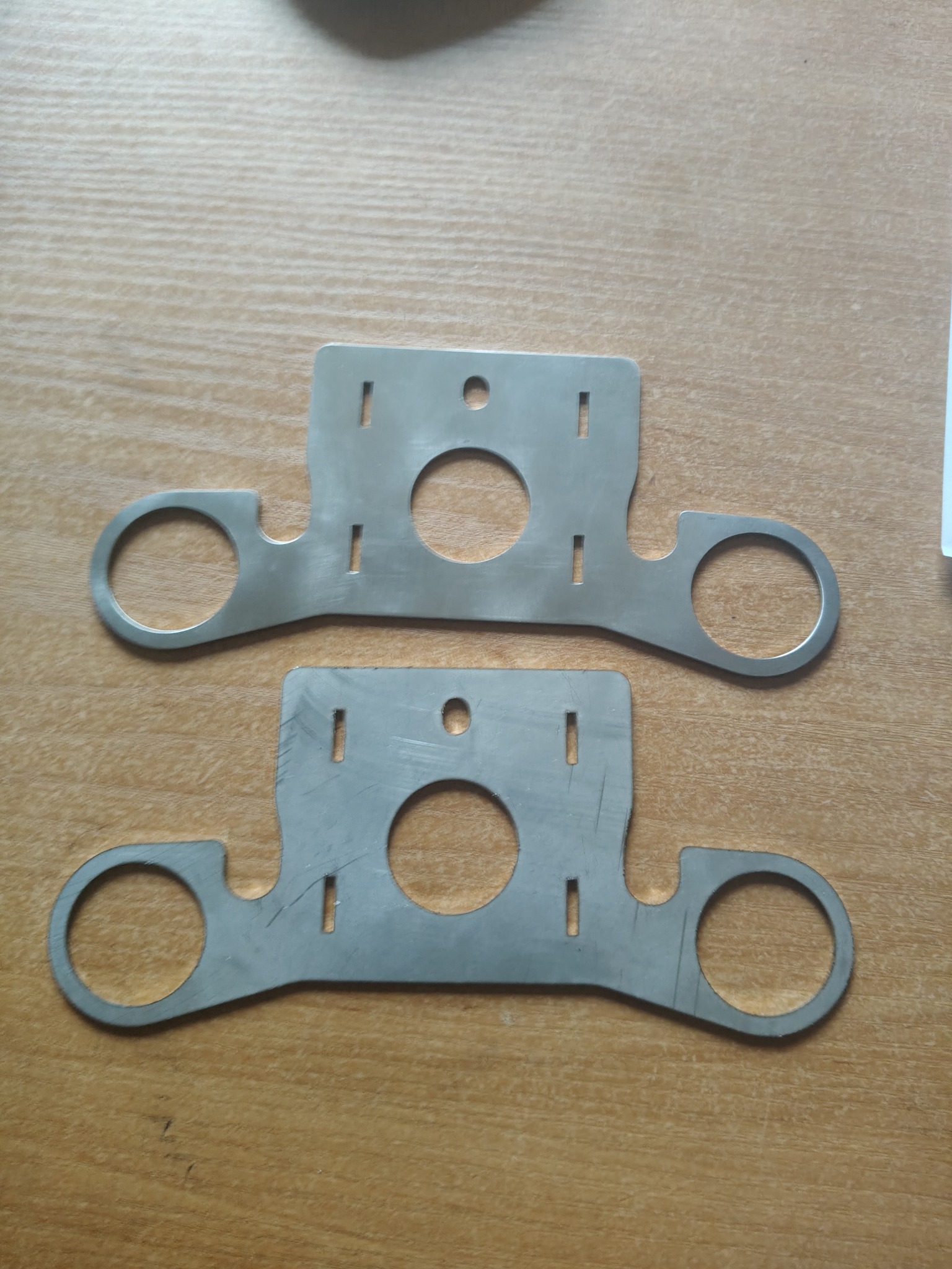 Dwie identyczne, płaskie, metalowe płytki z wyciętymi otworami (okrągły centralny otwór i dwa okrągłe otwory na końcach ramion) oraz podłużnymi nacięciami, leżące na drewnianym blacie.