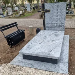 Jasnoszary, granitowy pomnik z krzyżem i wyrytym napisem, uzupełniony czarną, metalową ławeczką ze schowkiem na cmentarzu w Stargardzie.