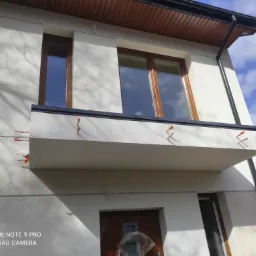 Elewacja budynku z balkonem w trakcie wykończenia, widoczne brązowe okna i pomarańczowe elementy mocujące na elewacji, błękitne niebo w tle.