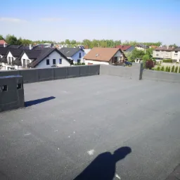 Widok z góry na płaski dach pokryty czarną papą, z widocznymi kominami wentylacyjnymi i zabudową attyki, w tle zabudowa jednorodzinna i zieleń.