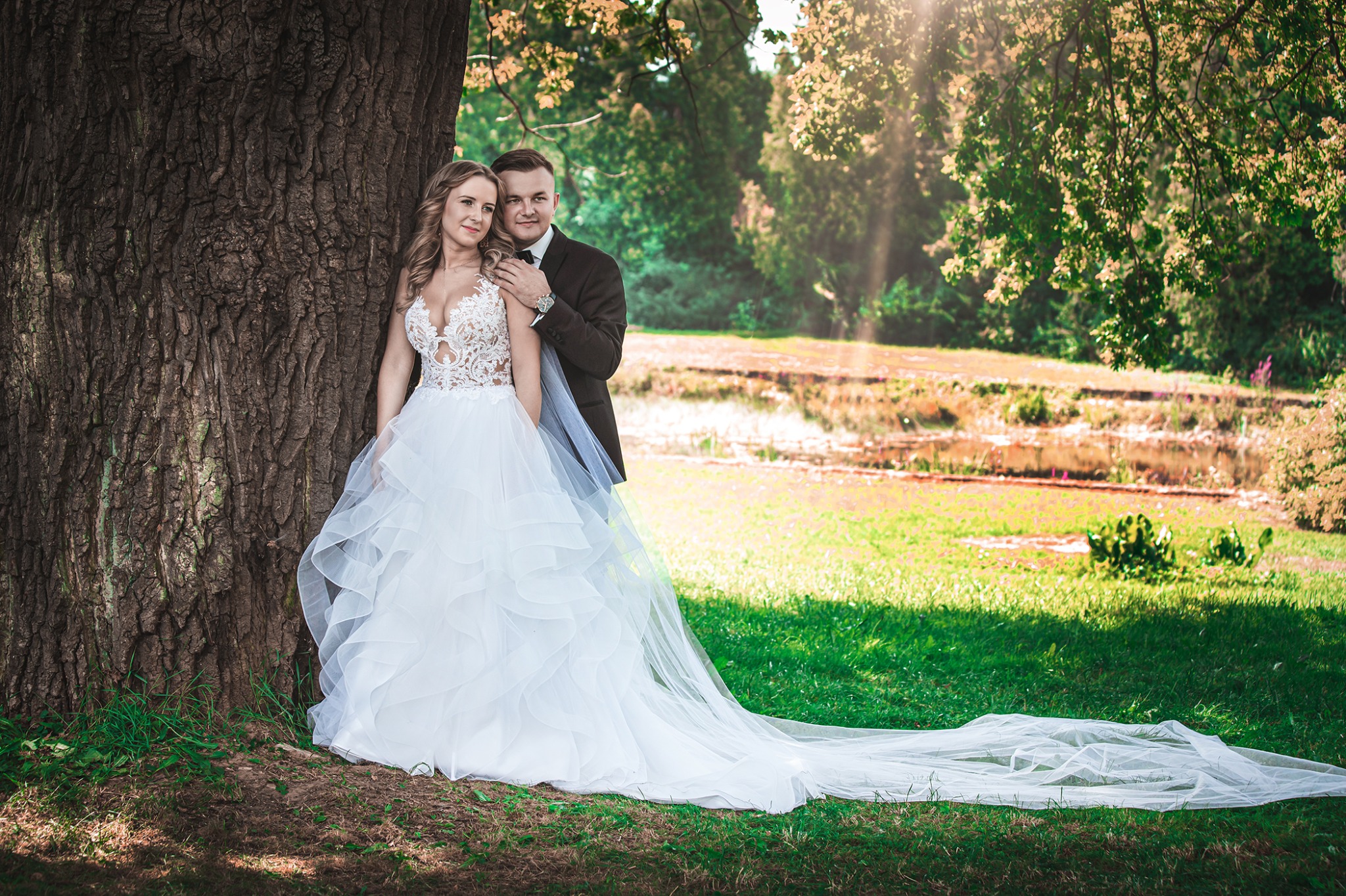 Para młoda, ona w koronkowej sukni ślubnej z długim trenem, on w ciemnym garniturze, pozują opierając się o stare drzewo w parku, w tle staw i promienie słońca.