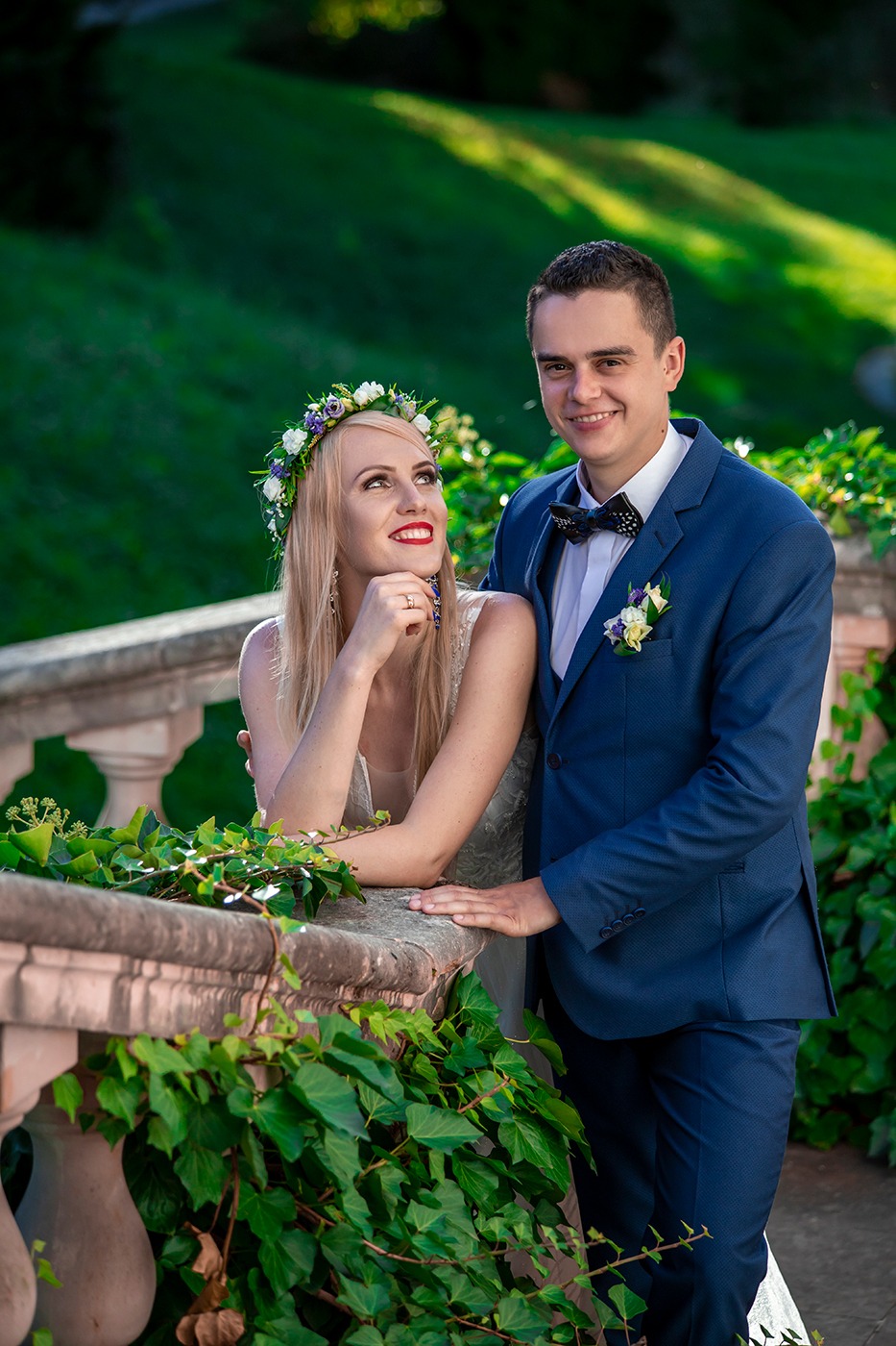 Uśmiechnięta para narzeczonych w eleganckich strojach pozuje na tle balustrady porośniętej bluszczem, panna młoda z wiankiem na głowie spogląda w górę.