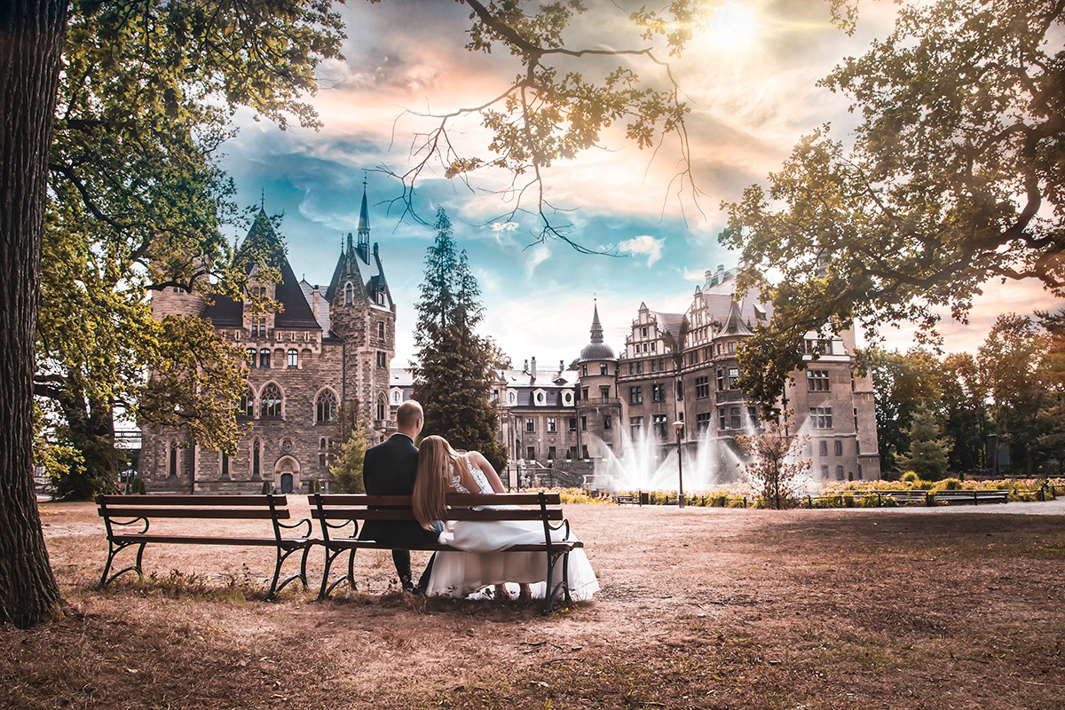 Para młoda siedzi na ławce, tyłem do widza, na tle zamku z fontannami; panna młoda ma długie, jasne włosy i białą suknię, pan młody w ciemnym garniturze.