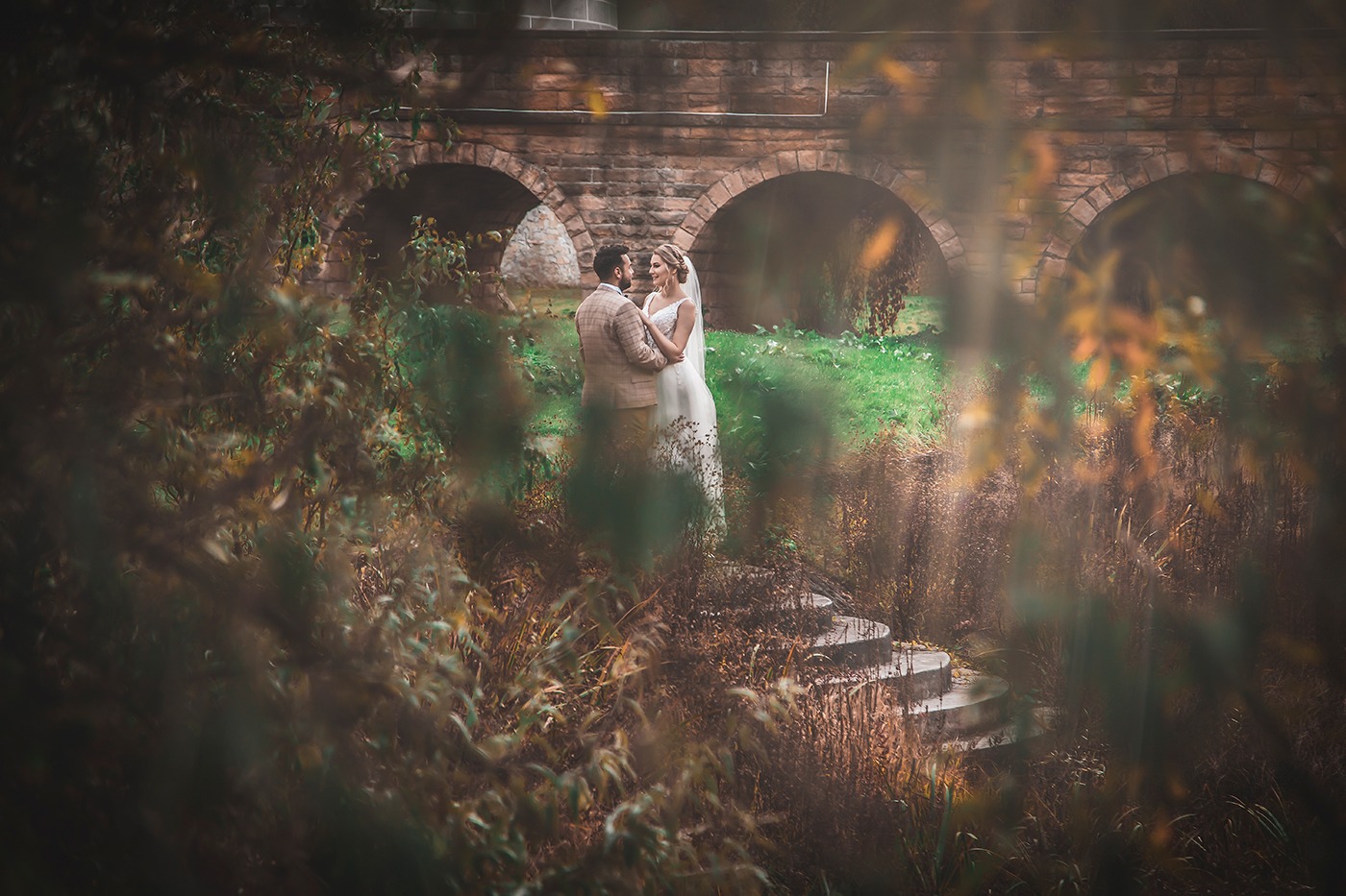 Para młoda obejmująca się na schodach przed kamiennym mostem z trzema łukami, widok przez zarośla na pierwszym planie.