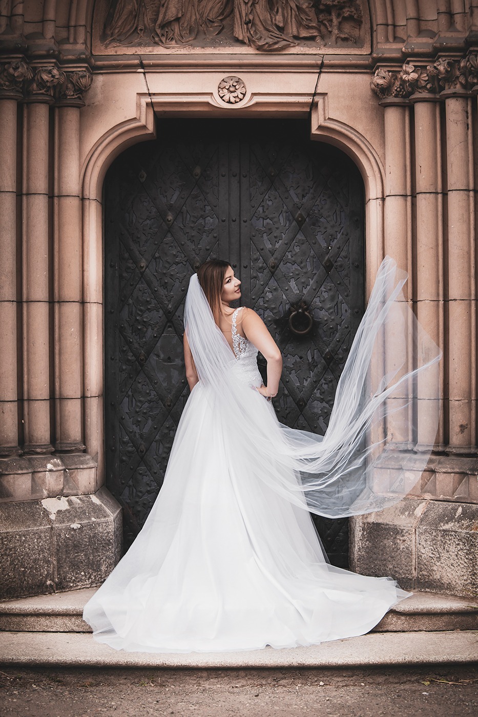 Panna młoda w białej sukni z koronkowym wykończeniem na plecach, stojąca tyłem przed ozdobnymi, ciemnymi drzwiami z kutego żelaza i kamiennym portalem, z welonem powiewającym na wietrze.