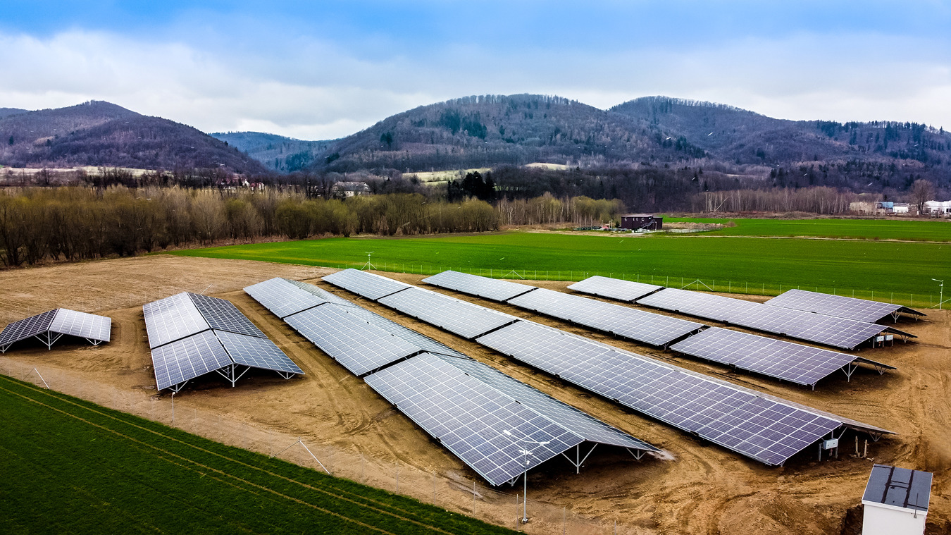 Rozległa farma paneli słonecznych w krajobrazie rolniczym z pagórkowatym tłem, widok z góry ukazuje rzędy paneli na brązowej ziemi, obok zielone pola i odległe budynki.
