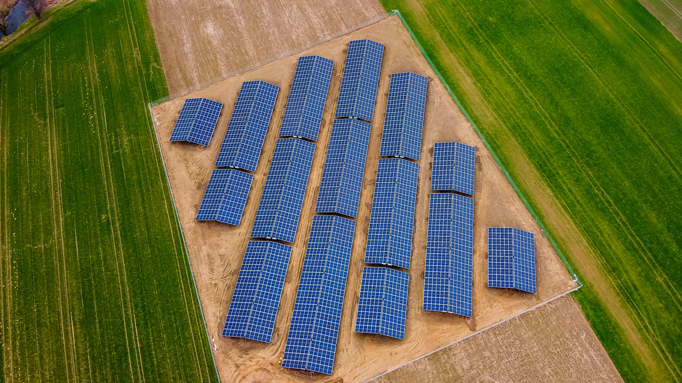 Widok z lotu ptaka na instalację paneli słonecznych na polu, z widoczną granicą między zieloną łąką a suchą ziemią, otoczoną siatką.