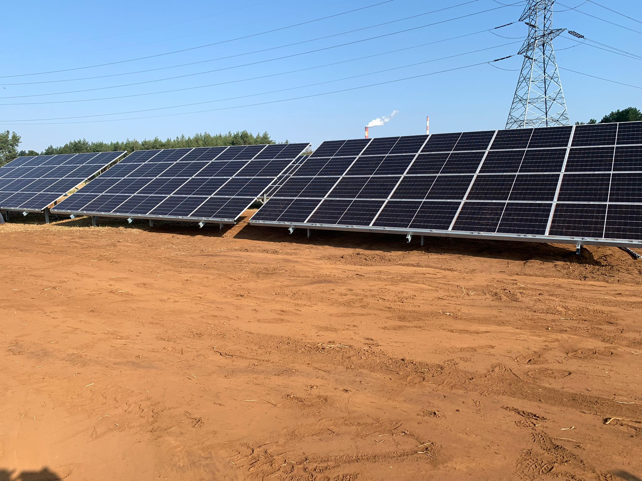 Rząd paneli słonecznych na brązowej ziemi, w tle słup wysokiego napięcia i komin z dymem, widok w słoneczny dzień.