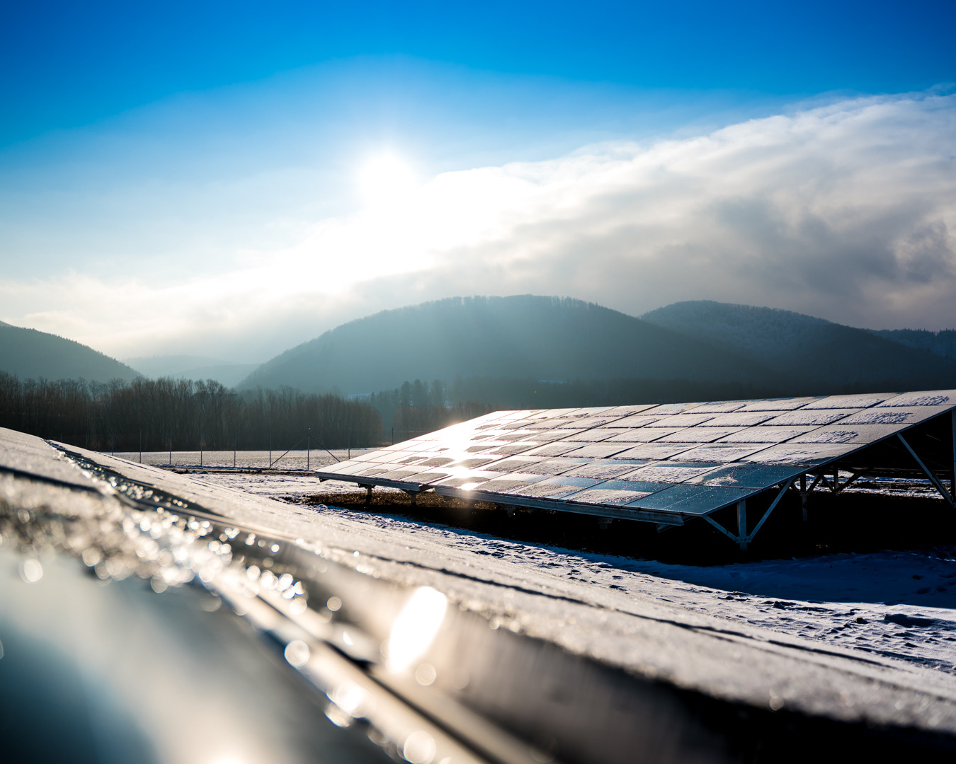 Zimowy krajobraz z panelami słonecznymi pokrytymi szronem, góry w tle, błękitne niebo z promieniami słońca odbijającymi się od powierzchni paneli.