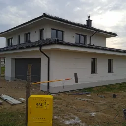 Nowoczesny dom jednorodzinny z szarą stolarką okienną, elewacja w trakcie malowania wałkiem na długim kiju, żółta skrzynka gazowa na pierwszym planie.