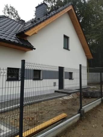 Nowo wybudowany dom jednorodzinny z białą elewacją, czarną dachówką i ogrodzeniem panelowym w trakcie montażu, widoczne niedokończone prace ziemne.