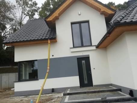 Nowoczesny dom jednorodzinny z białą elewacją, szarym akcentem kolorystycznym, czarnymi oknami i drzwiami, oraz ciemną dachówką. Widoczna żółta rura spustowa i schody wejściowe z szarego betonu.