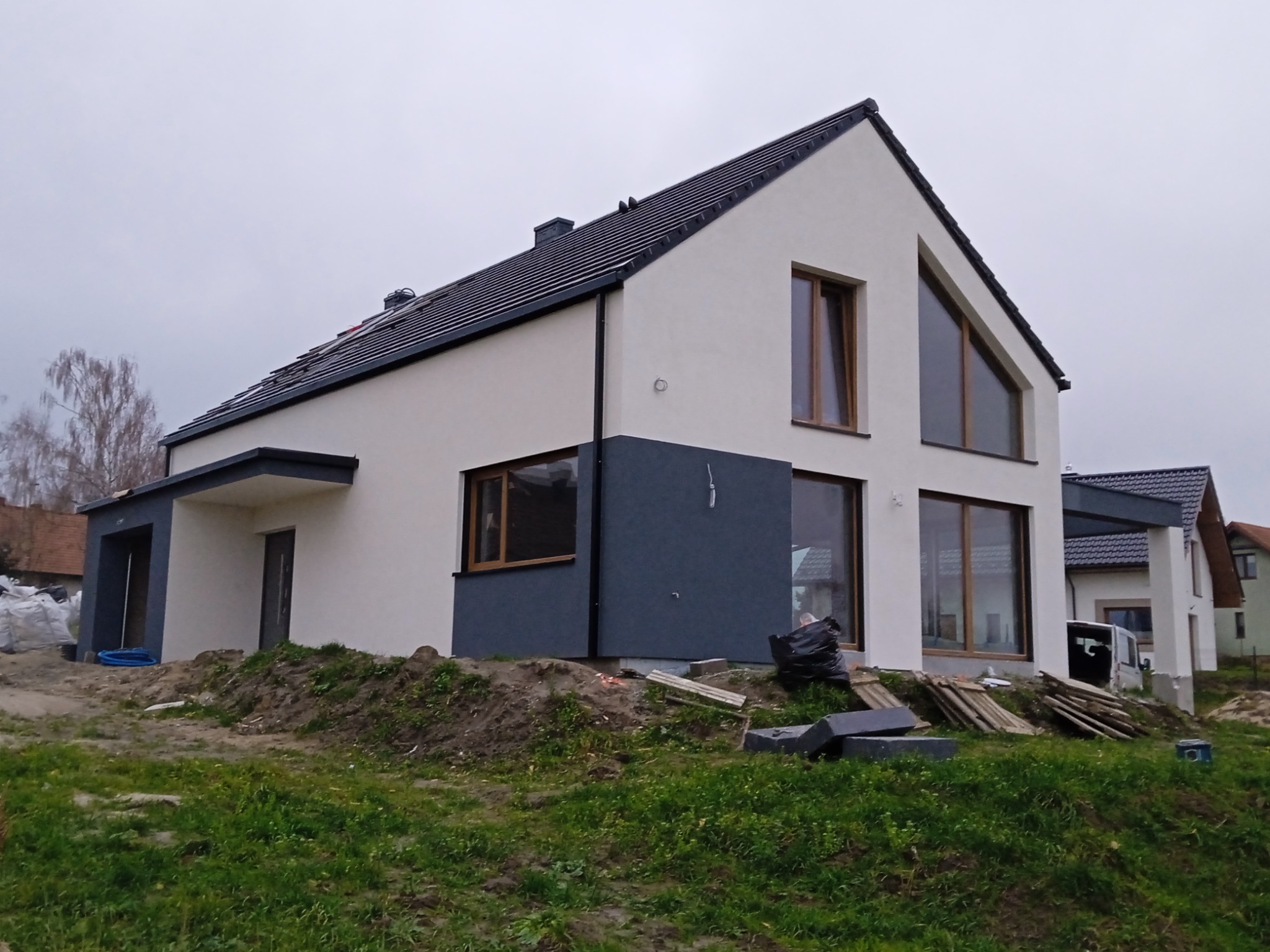 Nowoczesny dom jednorodzinny z białą elewacją i grafitowymi akcentami, widoczne prace wykończeniowe na zewnątrz, teren wokół domu częściowo uporządkowany, pochmurne niebo.