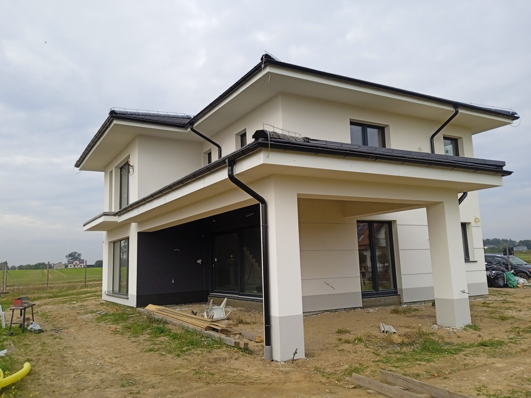 Nowoczesny, dwukondygnacyjny dom z płaskim dachem i wykończoną elewacją w odcieniach bieli i szarości, widok z zewnątrz na częściowo zagospodarowanym terenie budowy.