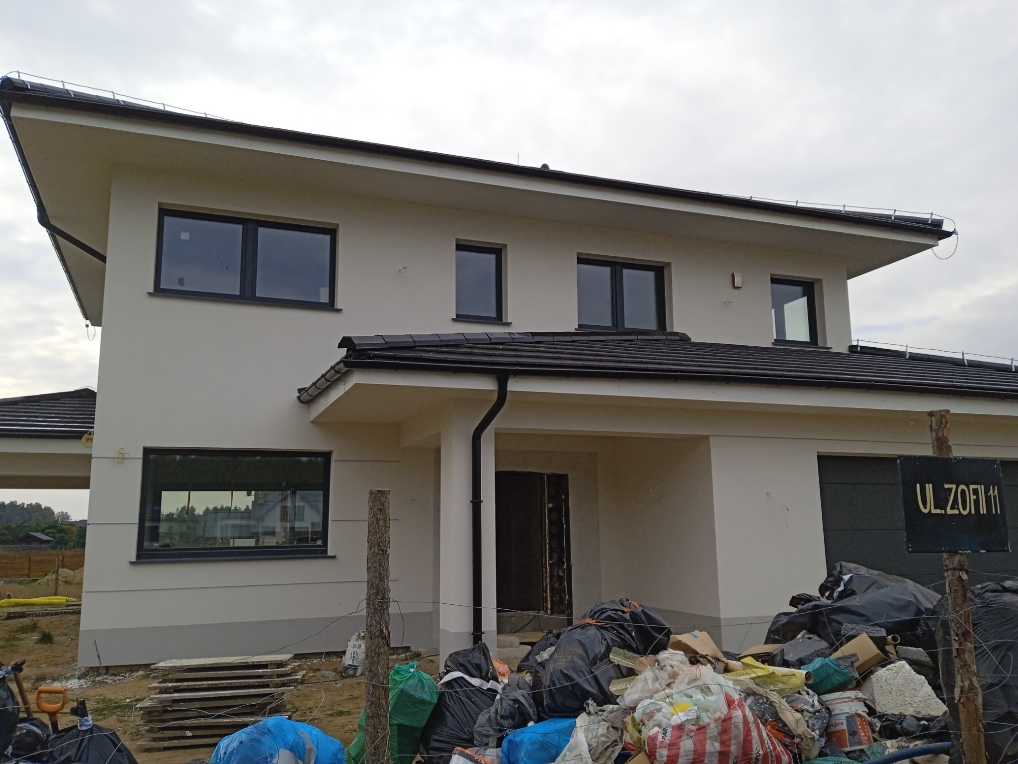 Nowoczesny dom jednorodzinny z płaskim dachem, świeżo po pracach wykończeniowych elewacji, z widocznymi czarnymi ramami okiennymi i stertą worków ze śmieciami budowlanymi przed budynkiem...