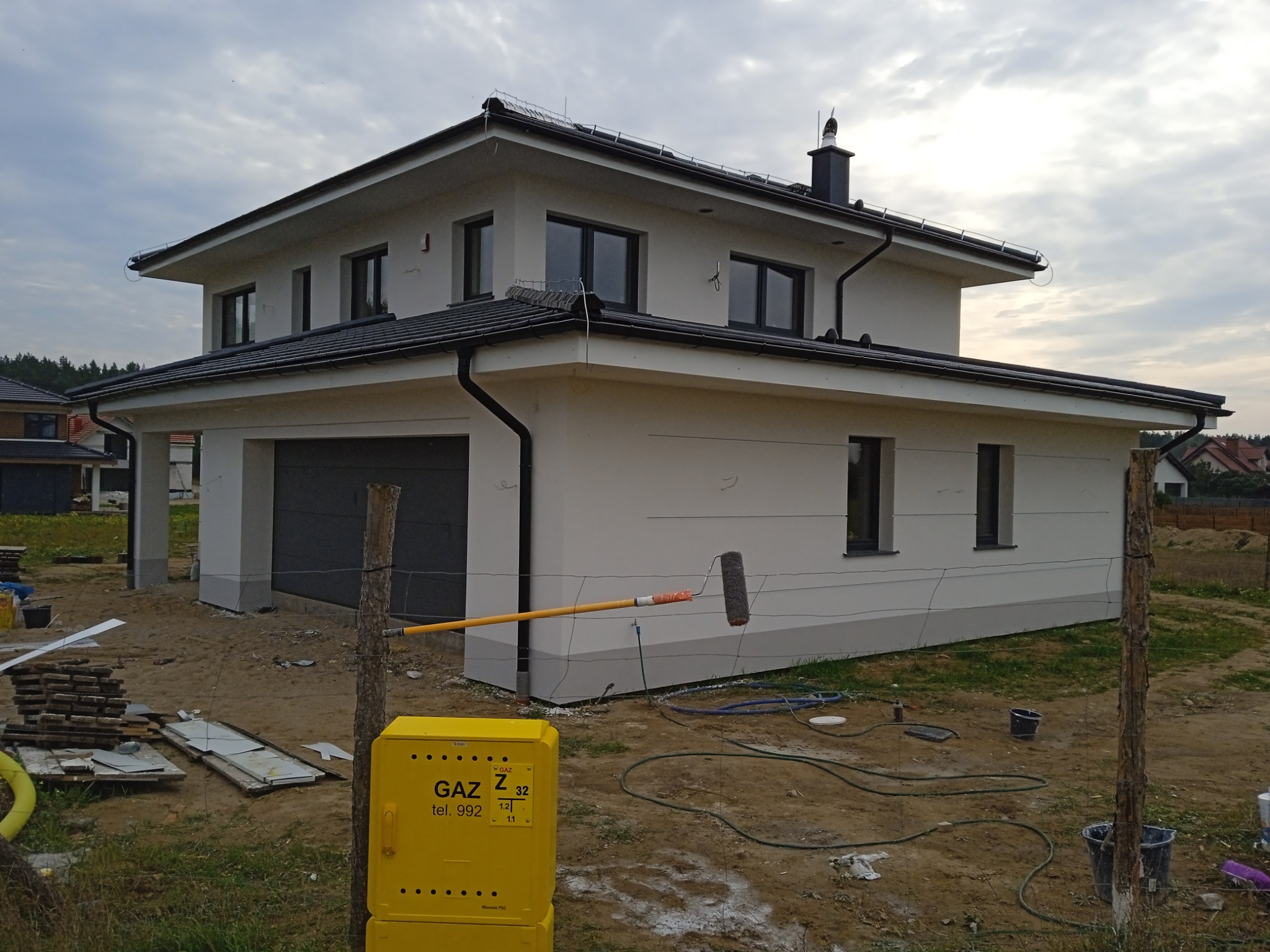 Nowoczesny dom jednorodzinny z szarą stolarką okienną, elewacja w trakcie malowania wałkiem na długim kiju, żółta skrzynka gazowa na pierwszym planie.