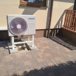 Zewnętrzna jednostka klimatyzacji Samsung zamontowana na metalowym stelażu przy ścianie budynku z widoczną kostką brukową.