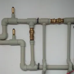 Szare rury instalacji wodnej z mosiężnymi zaworami i odpowietrznikami zamocowane na białej ścianie, widoczne obejmy mocujące.