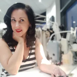 Portret kobiety z czerwonymi ustami w pracowni krawieckiej, w tle maszyna do szycia i wieszaki z ubraniami.