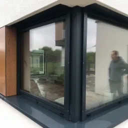 Nowoczesne okno narożne z ciemną ramą i drewnianą okładziną elewacyjną; odbicie sylwetki mężczyzny w szybie.