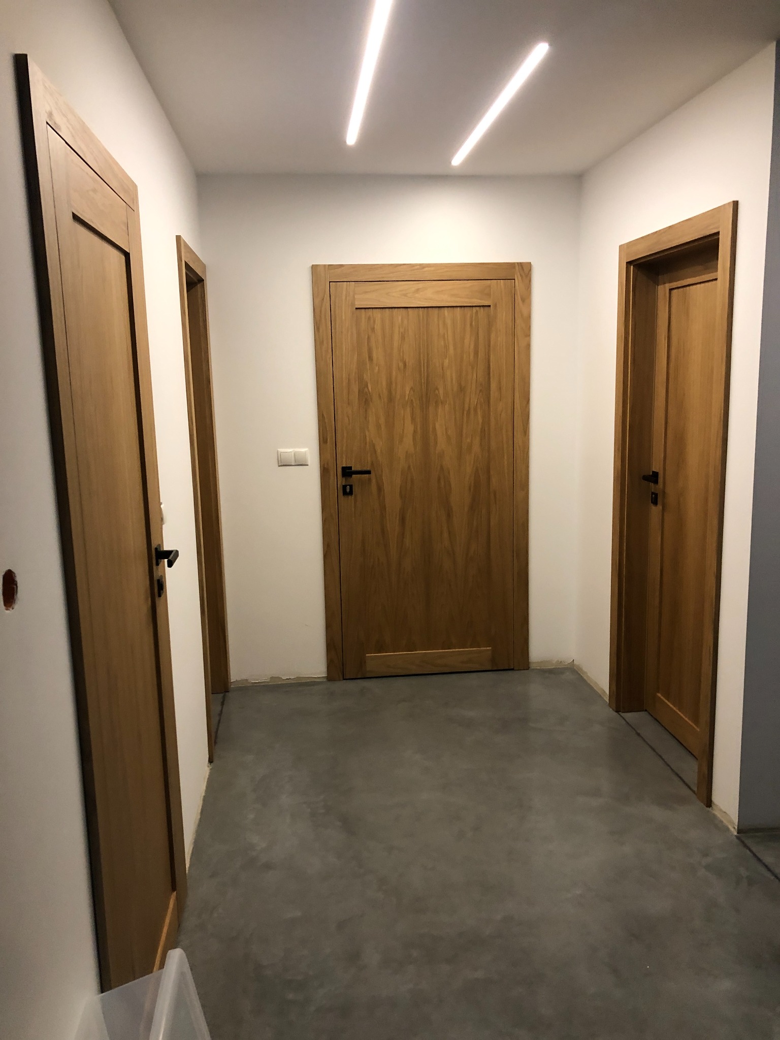 Wnętrze korytarza z trzema drewnianymi drzwiami z czarnymi klamkami, szarą posadzką i oświetleniem LED na suficie.