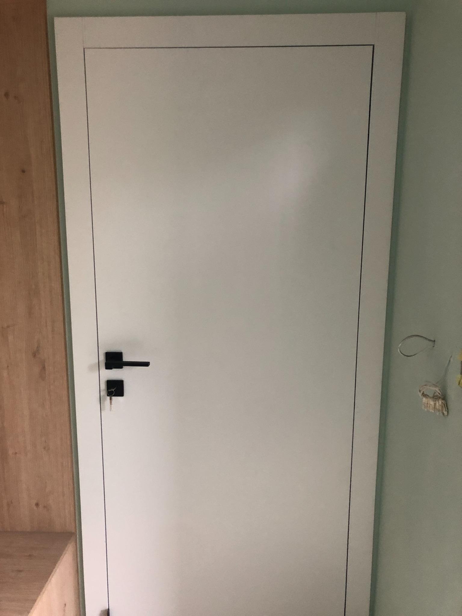 Zamknięte, białe drzwi wewnętrzne z czarną klamką i kluczem w zamku, widoczne na tle ściany w kolorze miętowym i fragmentu drewnianej zabudowy.