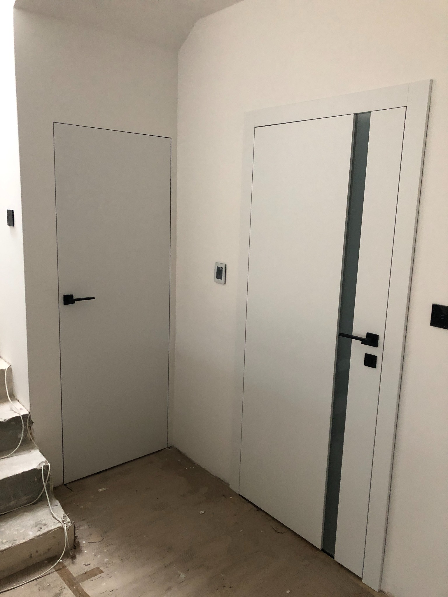 Dwa białe, minimalistyczne drzwi wewnętrzne z czarnymi klamkami w trakcie montażu, widoczne przewody elektryczne i niedokończone schody.