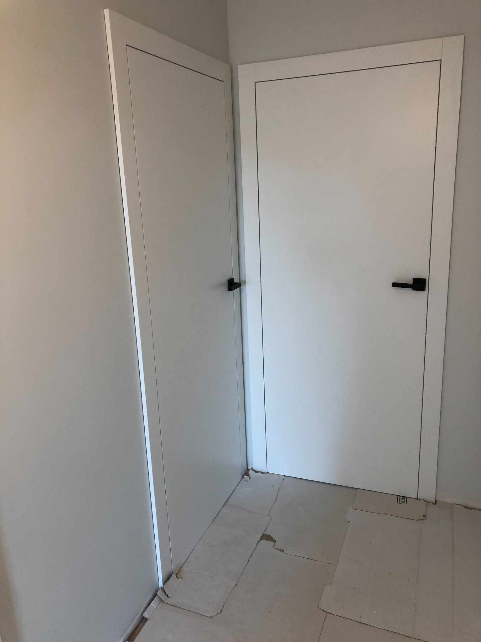 Dwa białe, minimalistyczne drzwi wewnętrzne z czarnymi klamkami na tle szarej ściany, częściowo zakryte kartonem zabezpieczającym podłogę.