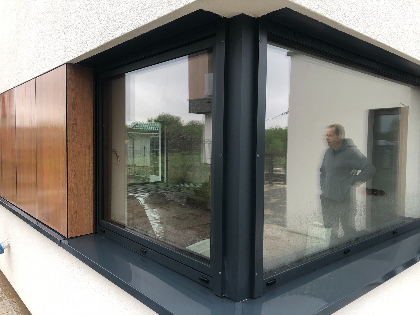 Nowoczesne okno narożne z ciemną ramą i drewnianą okładziną elewacyjną; odbicie sylwetki mężczyzny w szybie.