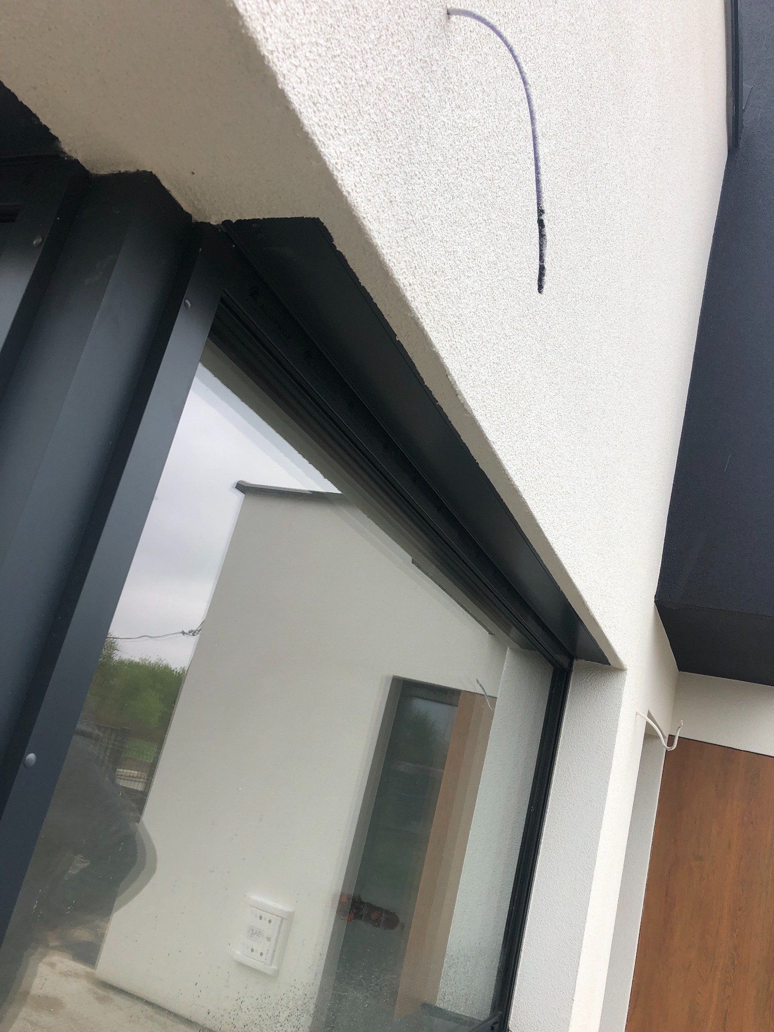 Zamknięte okno przesuwne z czarną ramą, widoczne z zewnątrz, z kablem wystającym ze ściany nad oknem i odbiciem budynku w szybie.
