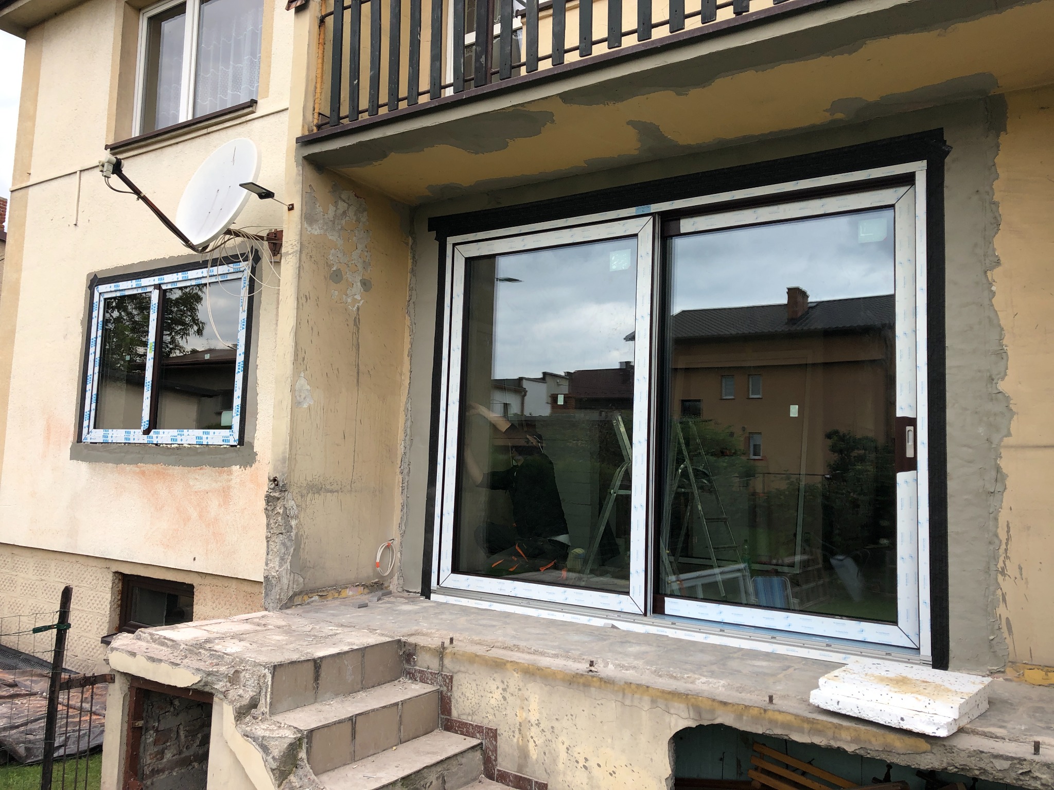 Nowe, przesuwne okno balkonowe z czarną ramą, zamontowane w surowym otworze, widoczne ślady prac murarskich i stare okno z anteną satelitarną obok.