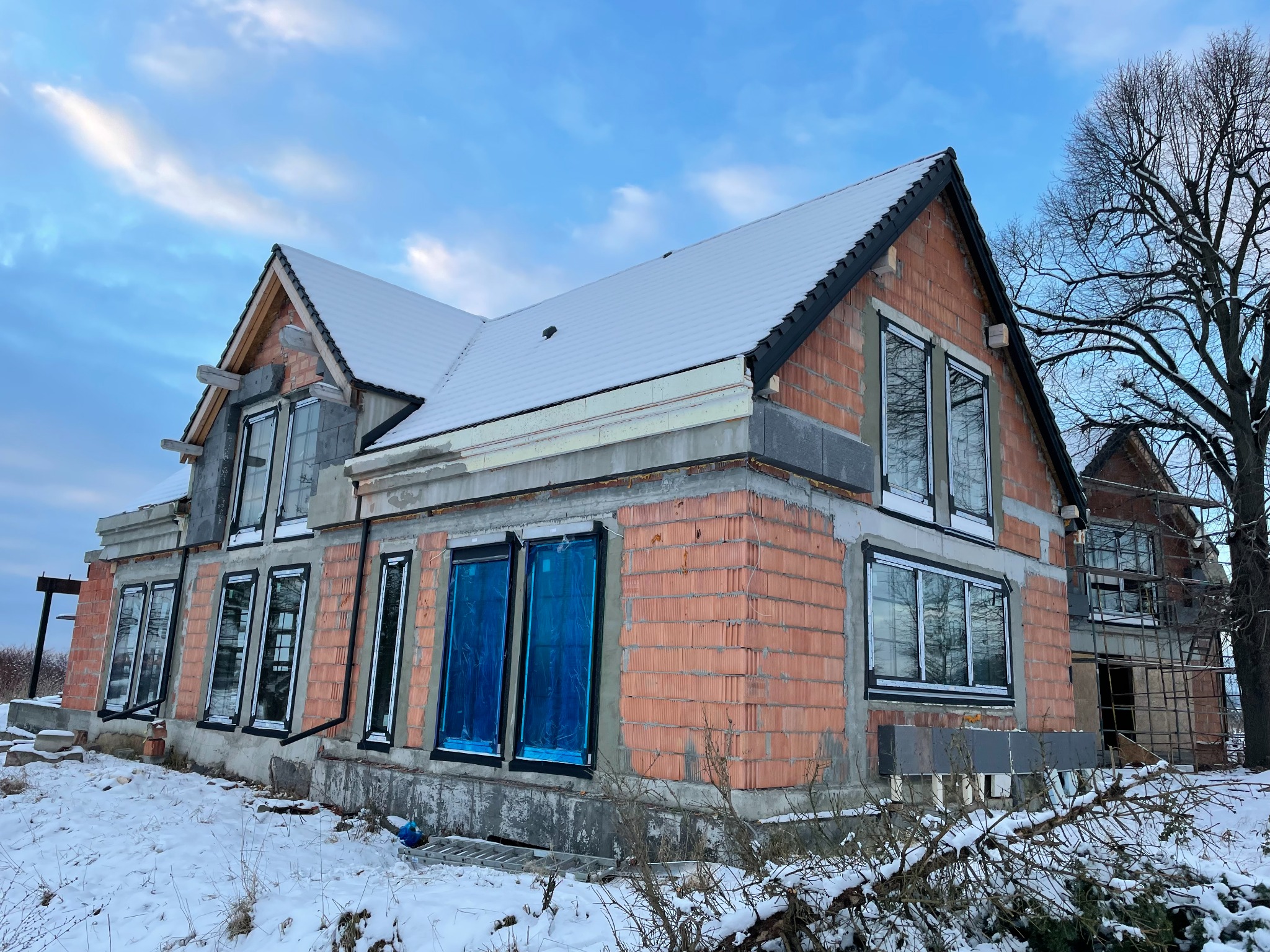 Niedokończony, dwupiętrowy dom z czerwonej cegły w zimowej scenerii, z częściowo zamontowanymi oknami zabezpieczonymi niebieską folią ochronną, pokryty śniegiem dach i widoczne rusztowanie z tyłu.