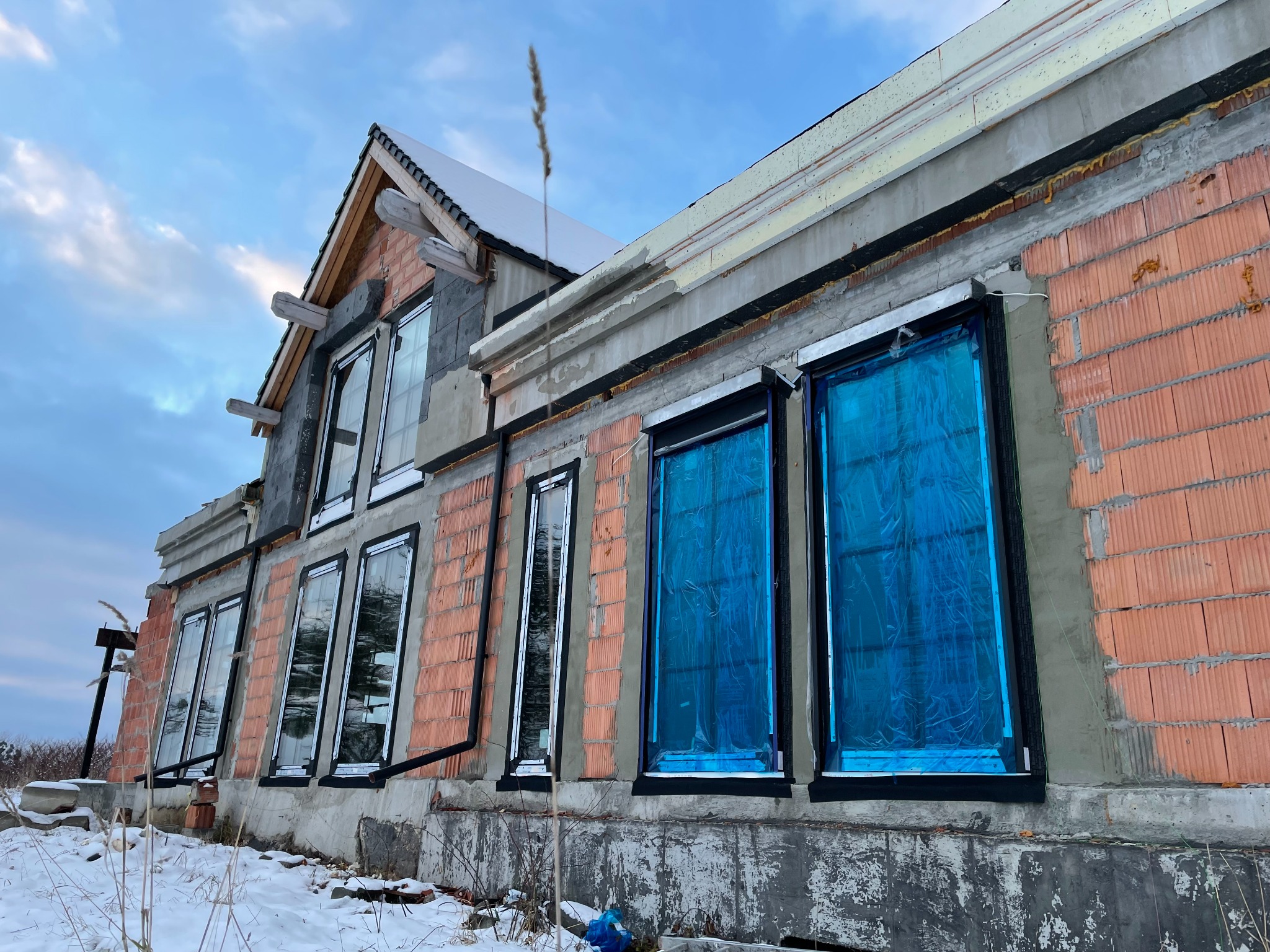 Surowy stan budynku w trakcie budowy z widocznymi oknami, z których część zabezpieczona jest niebieską folią, mur z cegły dziurawki, ośnieżone podłoże.