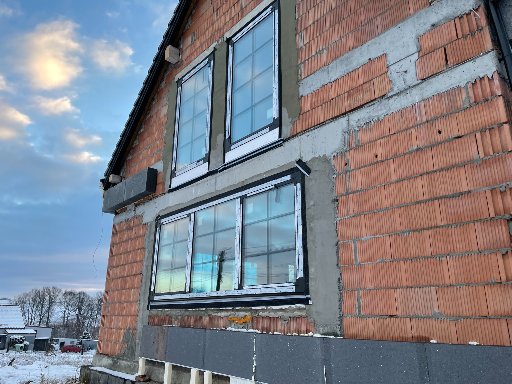 Surowy mur z czerwonej cegły porotherm z zamontowanymi oknami z PCV w czarnych ramach, widoczne ocieplenie budynku styropianem grafitowym, zimowy krajobraz w tle.