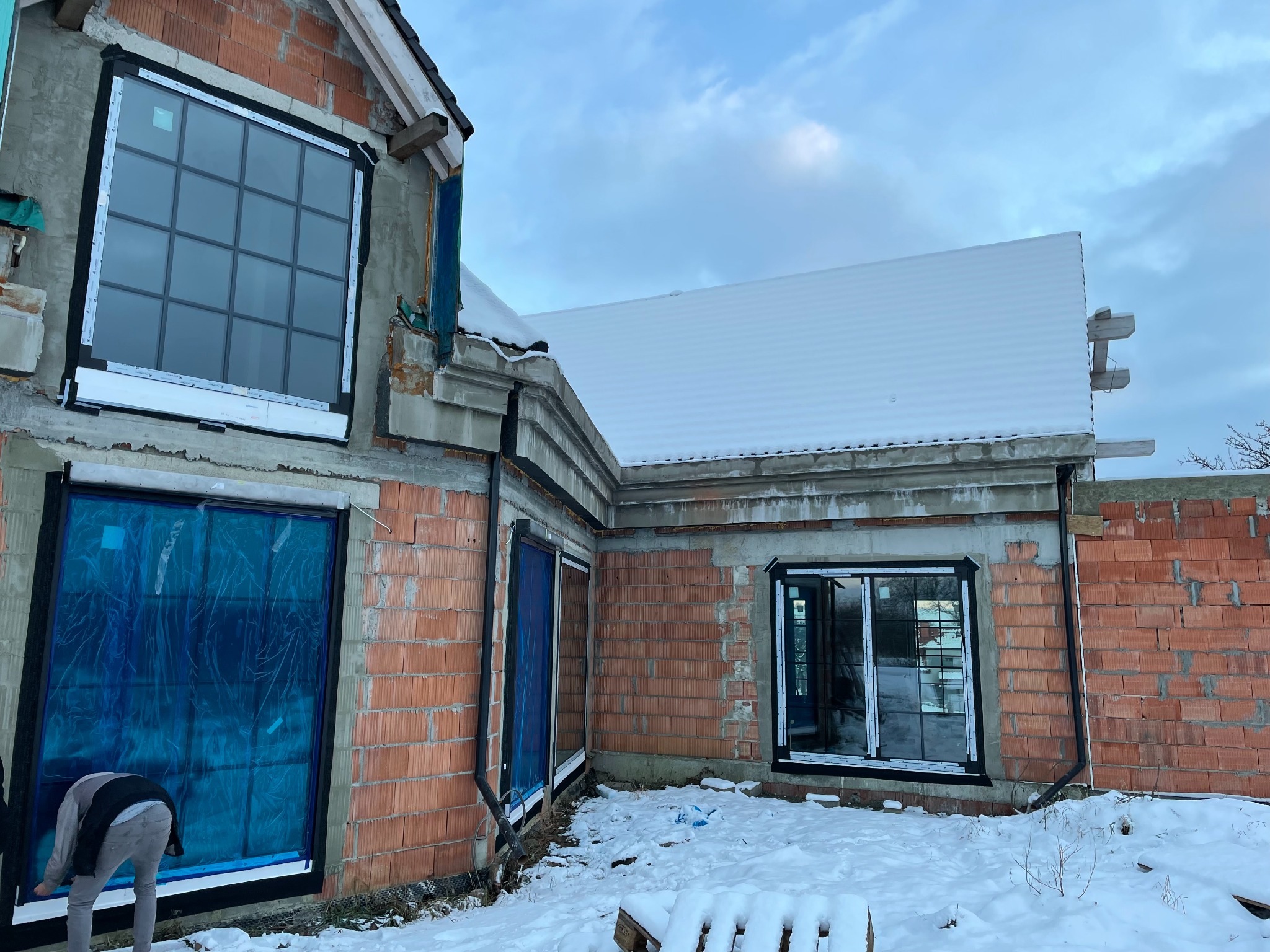 Montaż okien w surowym stanie budynku z czerwonej cegły w zimowej scenerii; okna zabezpieczone folią ochronną, widoczny pracownik przy oknie.