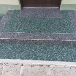 Schody wejściowe z żywicy epoksydowej w odcieniach zieleni i szarości prowadzące do brązowych drzwi z elementem dekoracyjnym, wykończone na zewnątrz budynku.