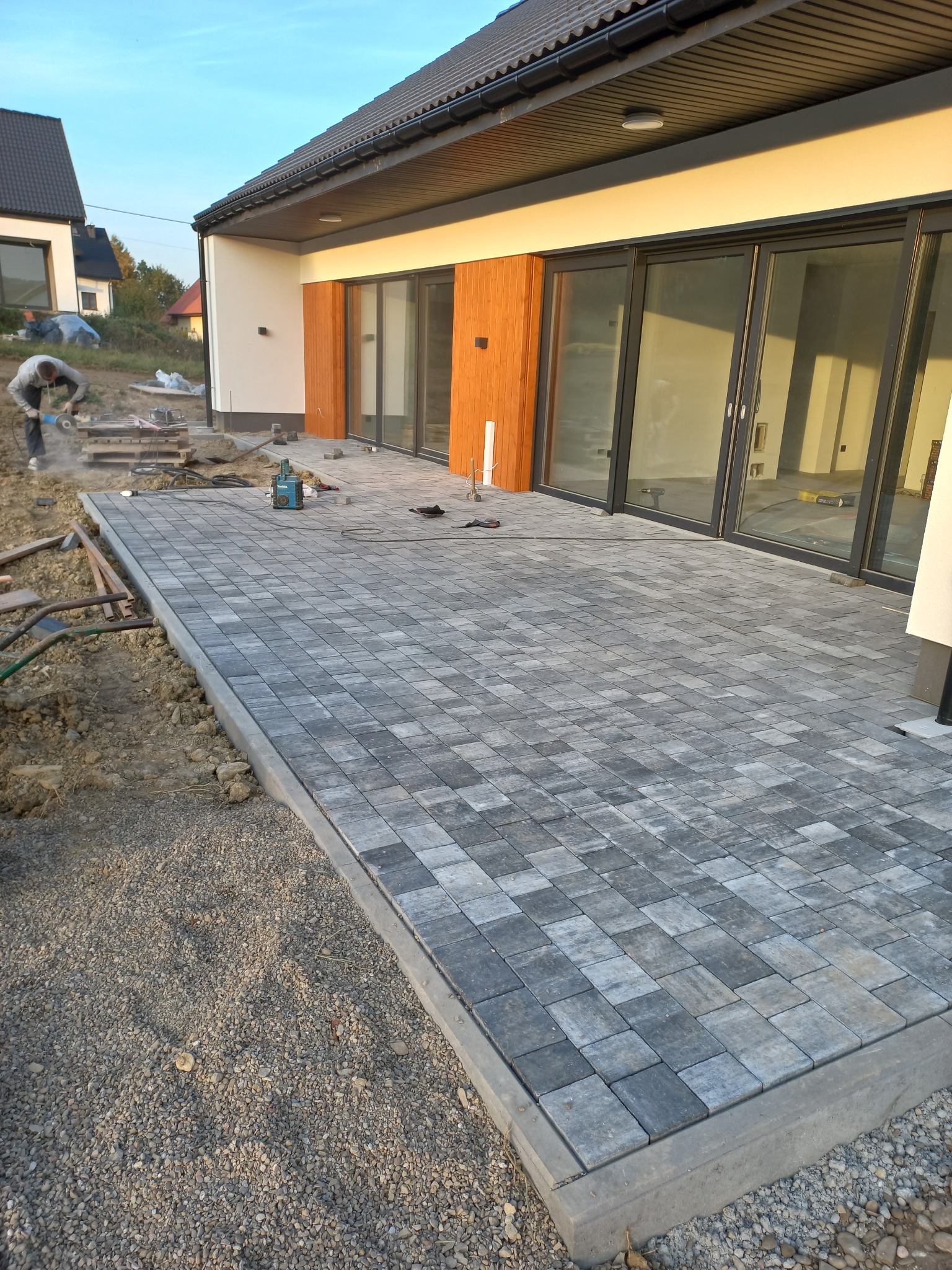 Układanie szarej kostki brukowej na tarasie przylegającym do domu z dużymi przesuwnymi oknami, widoczny pracownik przycinający kostkę szlifierką kątową.
