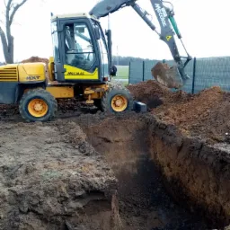 Żółta koparka Mecalac 12MXT w Rokosowie, w trakcie prac ziemnych związanych z kopaniem głębokiego wykopu, z widocznym fragmentem siatki ogrodzeniowej w tle.