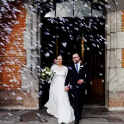 Para młoda wychodzi z kościoła podczas ceremonii ślubnej, obsypywana konfetti na tle ceglanej fasady i kamiennych łuków.